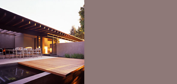Exterior view of a modern house with a wooden deck extending over a reflecting pool and a covered patio area with a pergola roof and metal b
