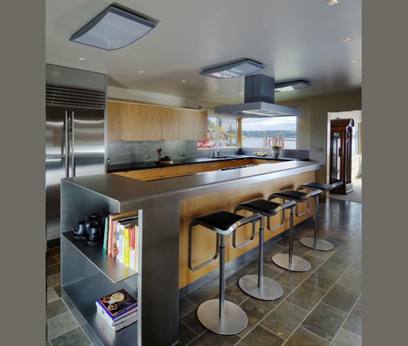 Modern kitchen with a center island featuring a metal countertop and wooden cabinetry, four bar stools, and stainless steel appliances.