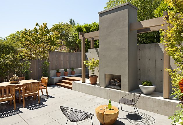 Sunlit outdoor living courtyard with a concrete fireplace and wooden dining set surrounded by greenery.