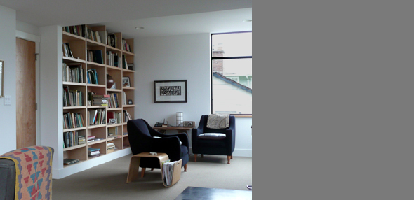 A bright living room features a built-in wooden bookshelf and two dark armchairs by a large window with black frames.