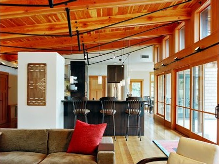 Interior view of a living room and kitchen with wood-paneled ceiling, large windows, and black bar stools at a kitchen island.