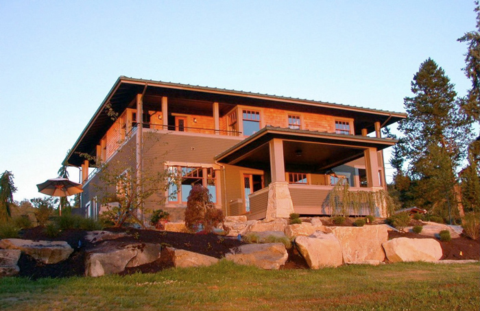 Two-story house with a large covered porch, wood siding, and stone foundation nestled against large rock landscaping.