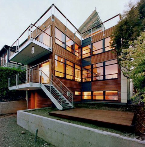 Modern multi-story house with wood siding, numerous large black-framed windows, and metal railings on the balconies and stairs at dusk.