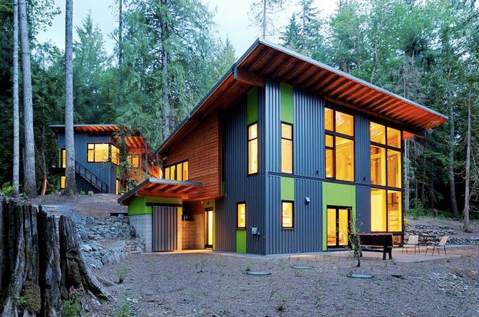 Modern two-story house with large windows and green and black siding surrounded by forest trees.
