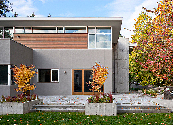 Front exterior of a modern two-story house with flat roof, concrete and wood siding, and fall-colored trees in planters.