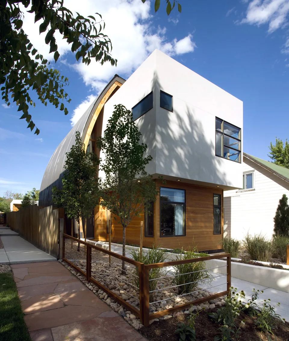 Exterior view of a modern two-story house with a white upper floor and wooden lower floor, featuring large windows and a curved metal roof,