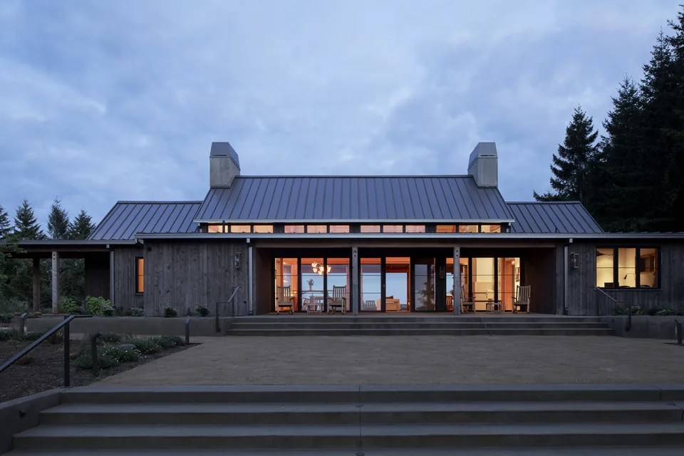 Exterior view of a modern farmhouse-style building at dusk with a metal gable roof, two chimneys, and large glass sliding doors opening to a