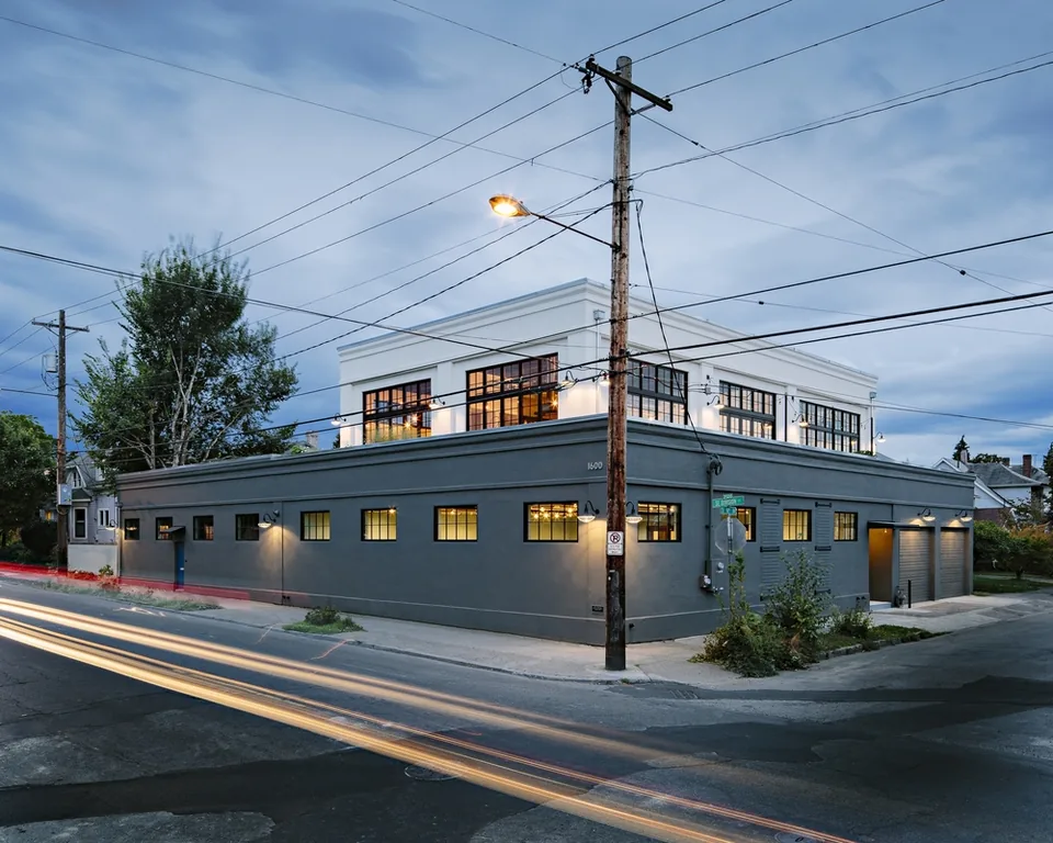Street view of a two-story modern building with large black-framed windows and exterior lighting at dusk.