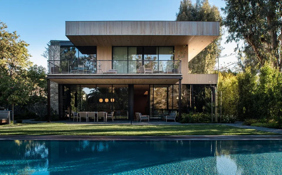Modern two-story house with wood panel exterior, large glass sliding doors, balcony with metal railing, and a swimming pool in the yard.