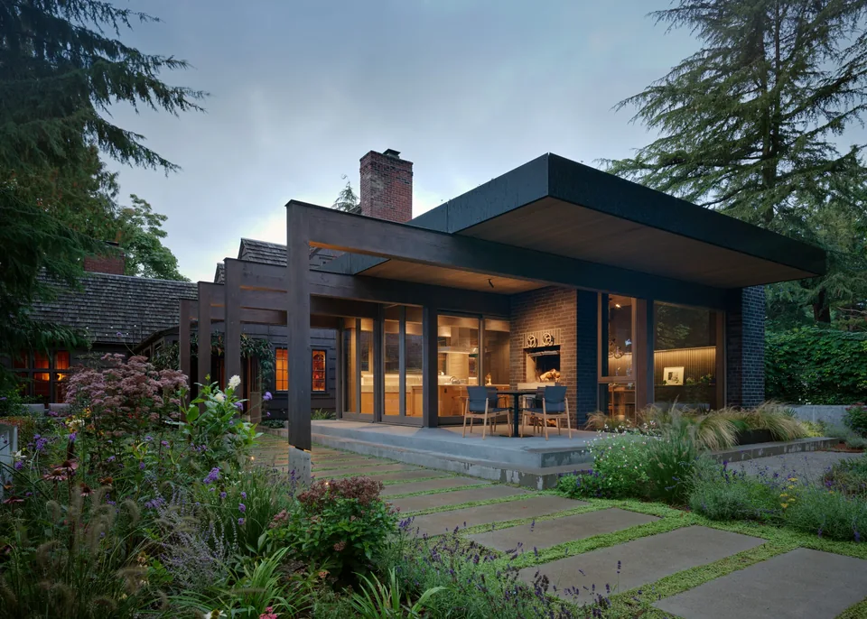 Modern house exterior featuring a covered patio with wood ceiling and black framing, with a brick chimney and large glass doors opening to a
