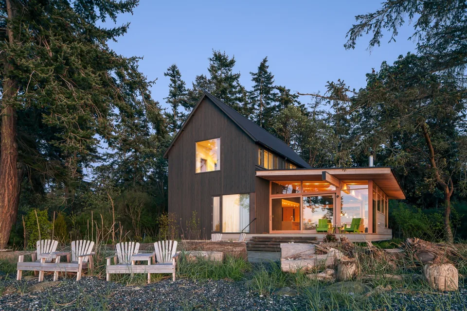 A modern two-story house with a steep gable roof and a flat-roofed extension surrounded by tall trees, with a seating area of wooden chairs.