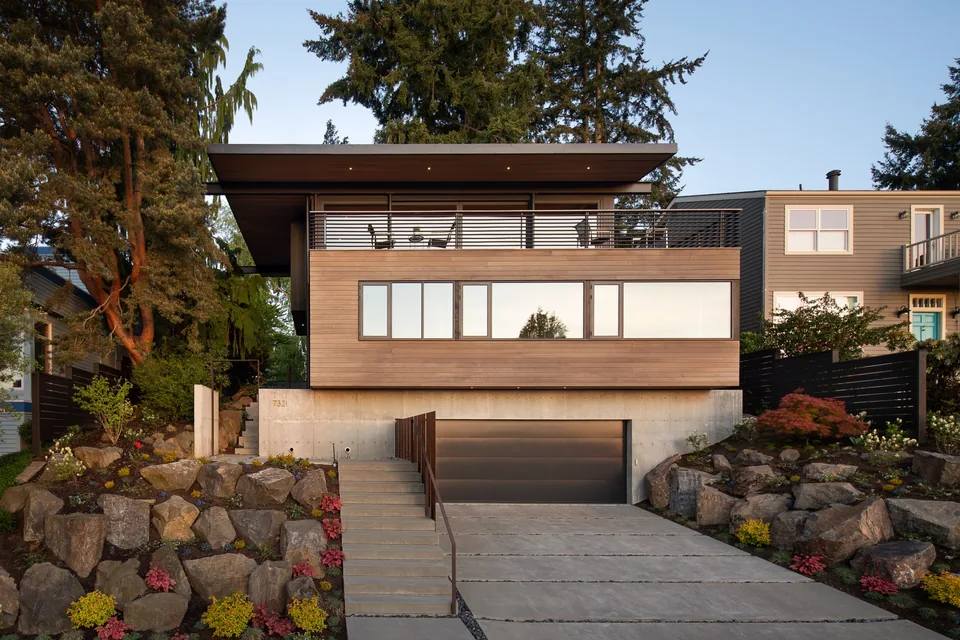 Modern two-story house on a raised cement foundation with horizontal wood siding and a flat roof, featuring large windows and a balcony with