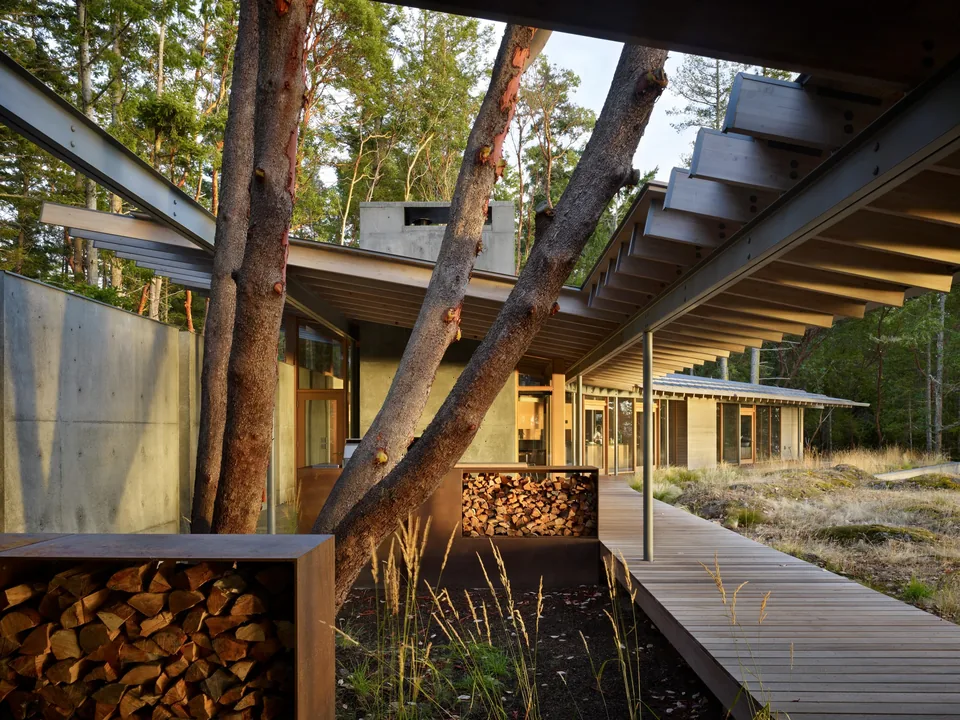 Exterior view of a modern house with exposed wooden beams and steel supports surrounding trees, featuring wood storage and a concrete wall,,
