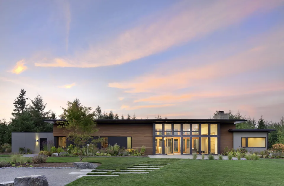 Single-story modern residence with wood siding and large floor-to-ceiling windows at dusk with landscaped front yard and stepping stone path