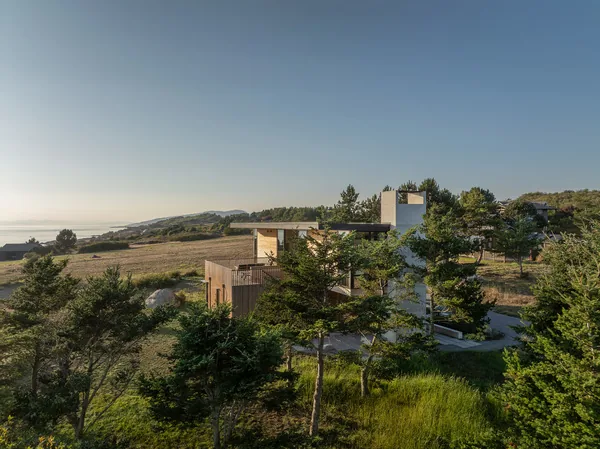 Modern house situated in a natural landscape with trees and grassy fields near a body of water under a clear sky.