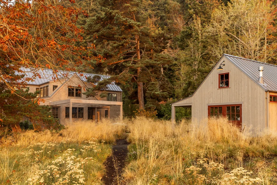Two light-colored wood houses with metal roofs surrounded by tall grasses and trees in a natural setting.