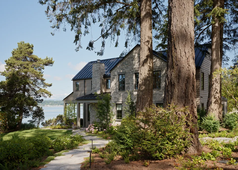Stone and wood two-story house with a metal roof surrounded by large trees and greenery overlooking a body of water.