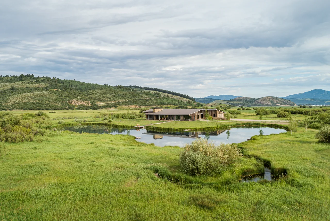 A low, single-story retreat house with wood siding and stone chimney reflects in a pond surrounded by green fields and distant hills.