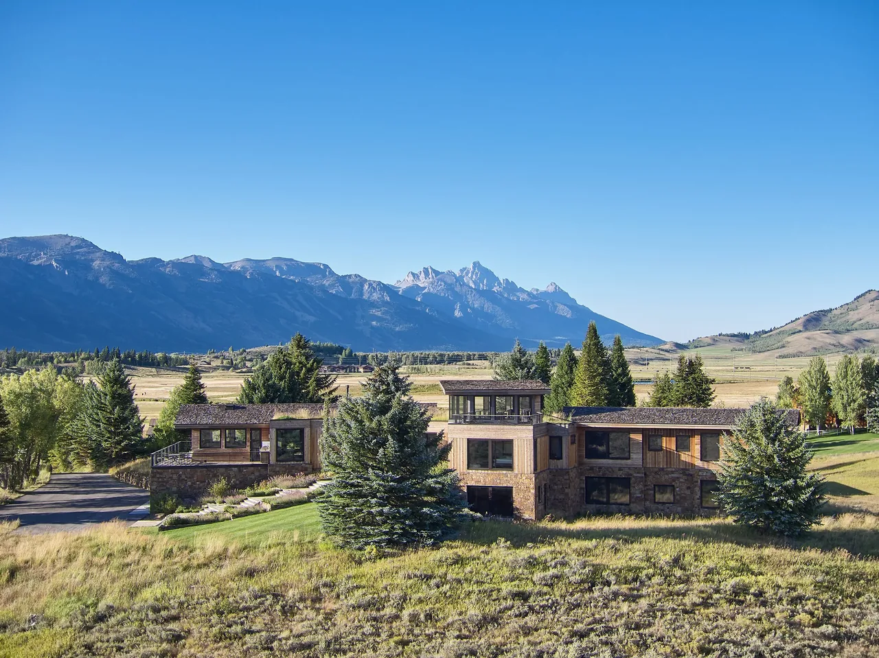 Wide exterior view of a multi-level residence with wood and stone cladding, set in a grassy landscape with mountains in the background.