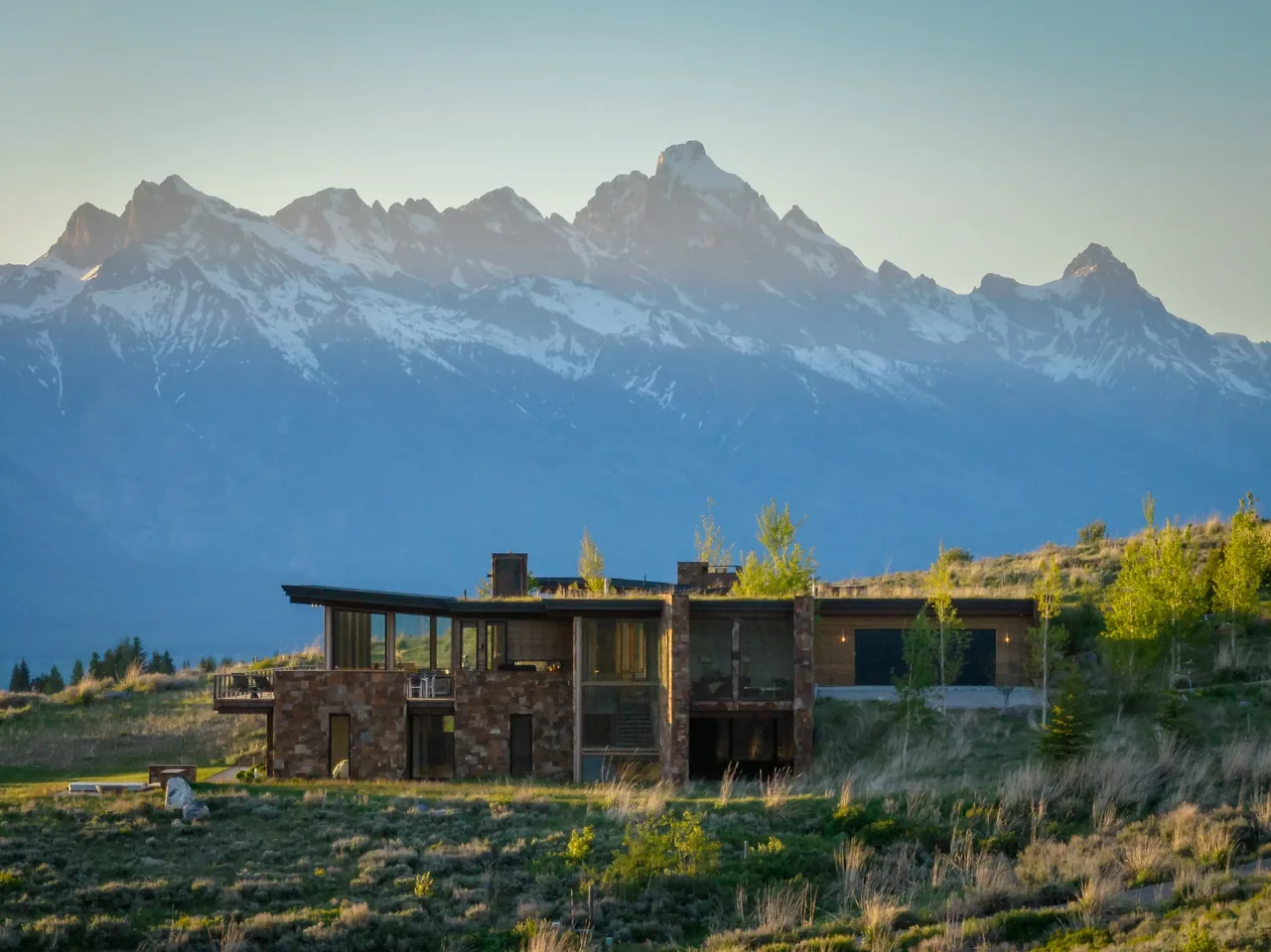 Modern residence with stone and wood exterior set against a backdrop of snow-capped mountains under a clear sky.
