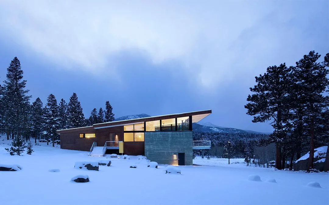 Modern house with slanted roof in a snowy landscape, surrounded by pine trees and mountains during dusk.