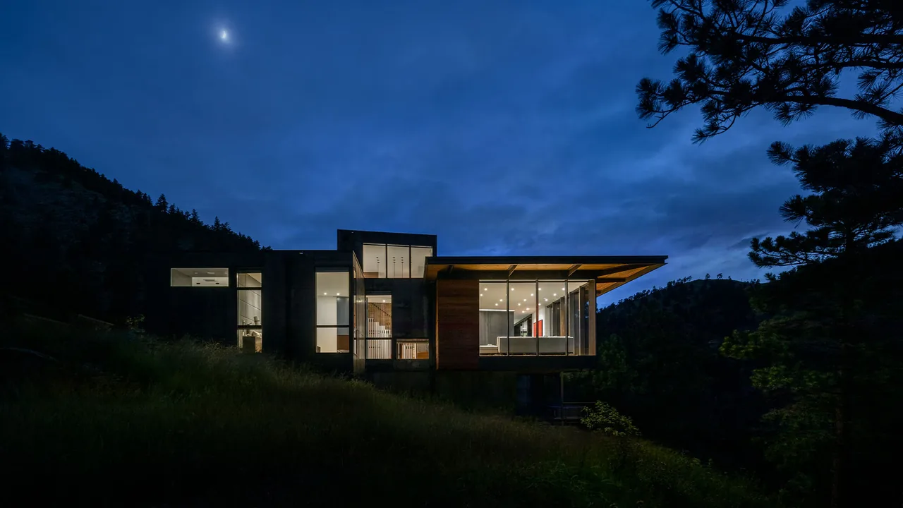 Night view of a modern house on a hillside with an illuminated interior and large floor-to-ceiling windows under a partly cloudy sky with a