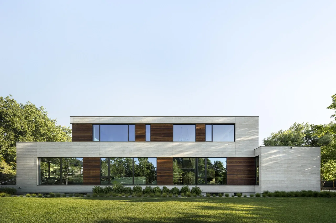 Modern two-story house exterior with large windows and sections of wood paneling, surrounded by trees and a lawn.