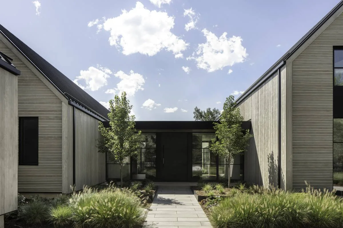 View of a modern residential entrance with concrete walkway, wood siding, and tall narrow windows under a partly cloudy sky.