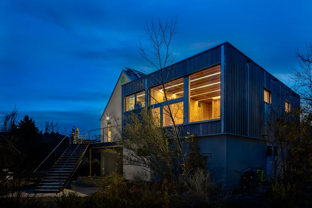 Modern two-story house at dusk with illuminated interior, featuring metal siding on the upper level and a staircase leading to an exterior,