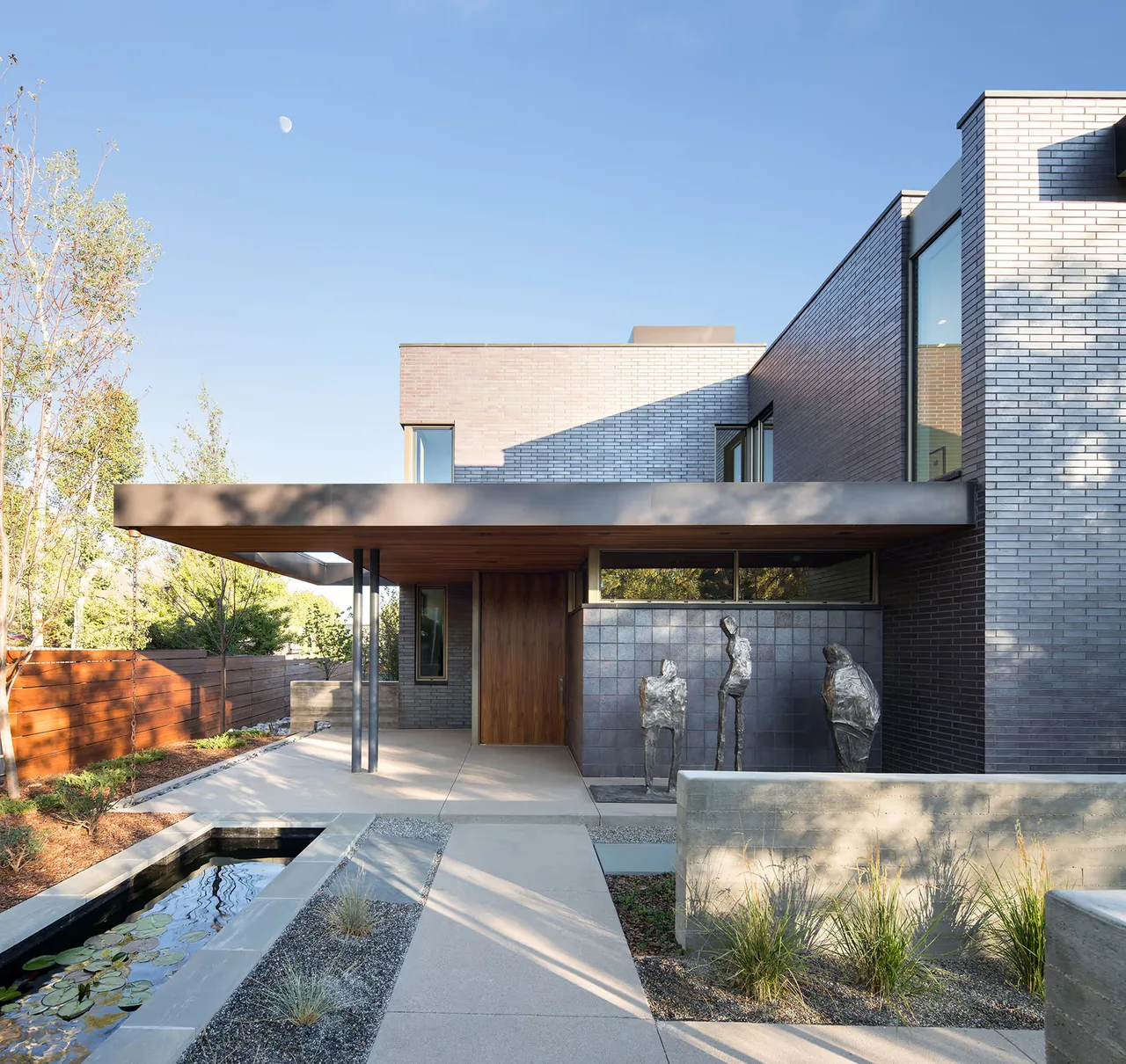 Modern two-story brick house with a flat roof, cantilevered entry canopy, and a reflecting water feature alongside the walkway.