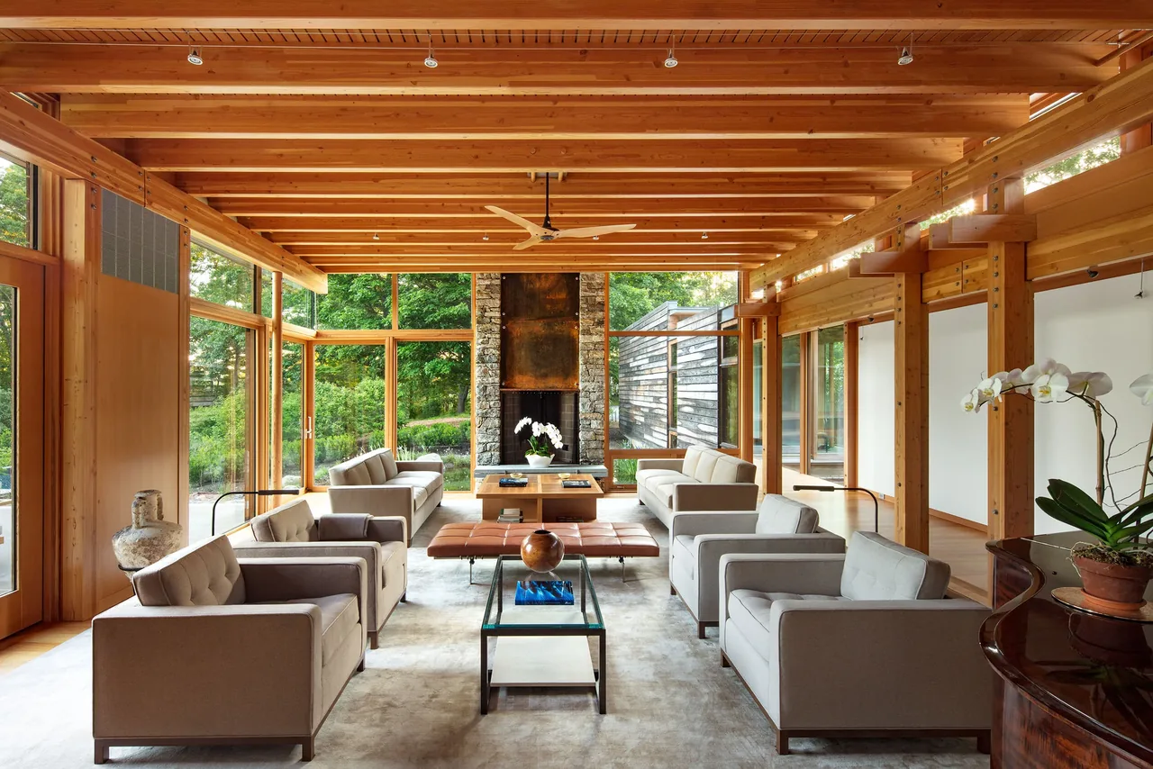 Spacious living room with exposed wood beams, large stone fireplace, and floor to ceiling windows overlooking greenery.