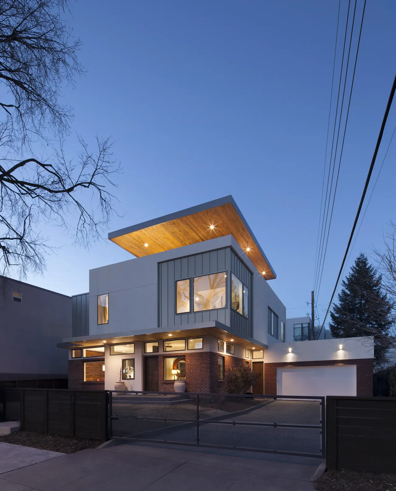 Modern two-story house with angled roof overhang, mixed cladding of brick, stucco, and metal paneling, illuminated at dusk.