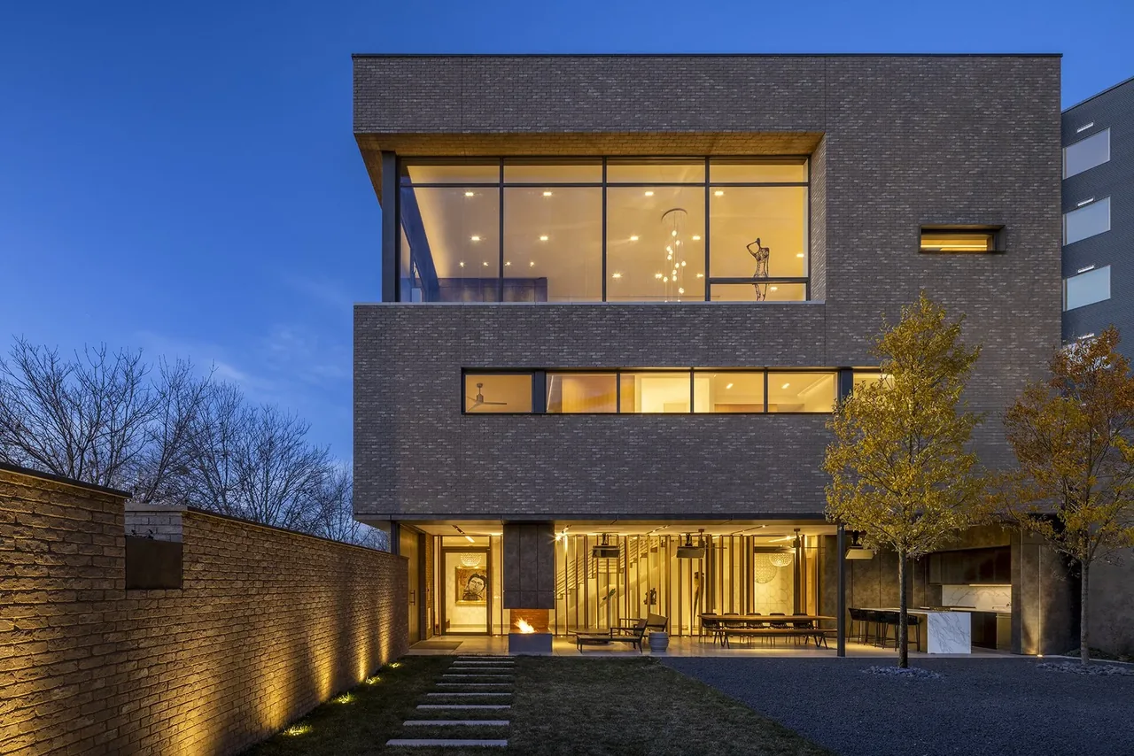 Modern three-story private residence with large floor-to-ceiling windows, brick exterior, lit at dusk with a landscaped yard and stone path.