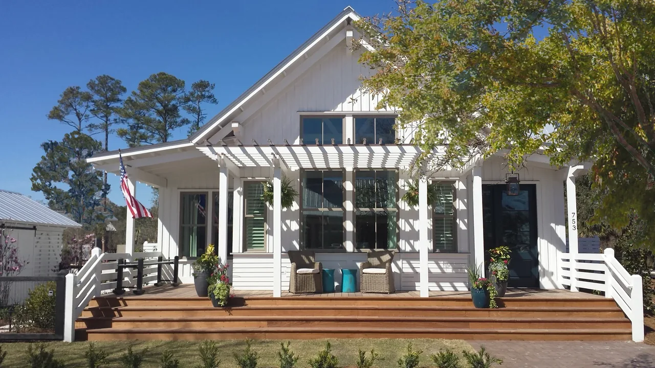 Front exterior of a white cottage with a gable roof, wooden deck, pergola, and railing in a landscaped yard.