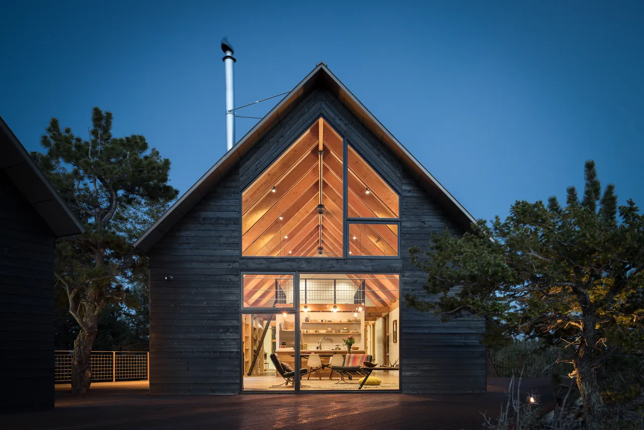 Night view of a black wood-sided cabin with large triangular and rectangular windows revealing a warmly lit interior with exposed wood beams
