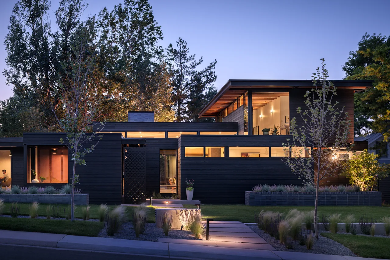 Modern two-story house with black wood siding and large horizontal windows illuminated at dusk.