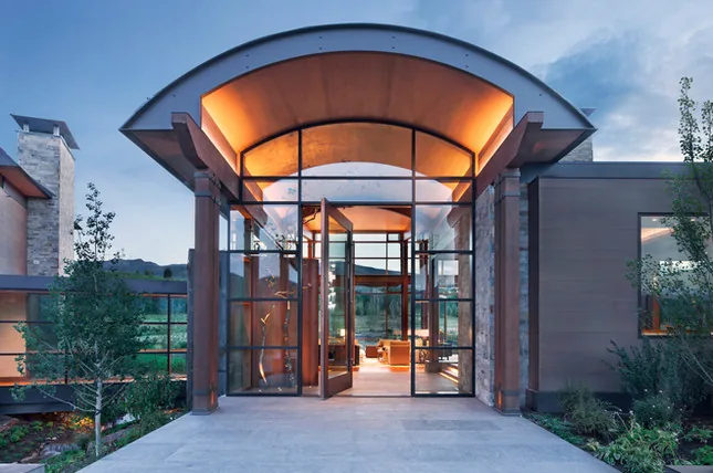 Exterior view of a modern house entrance with a curved roof and large floor-to-ceiling glass panels at dusk.