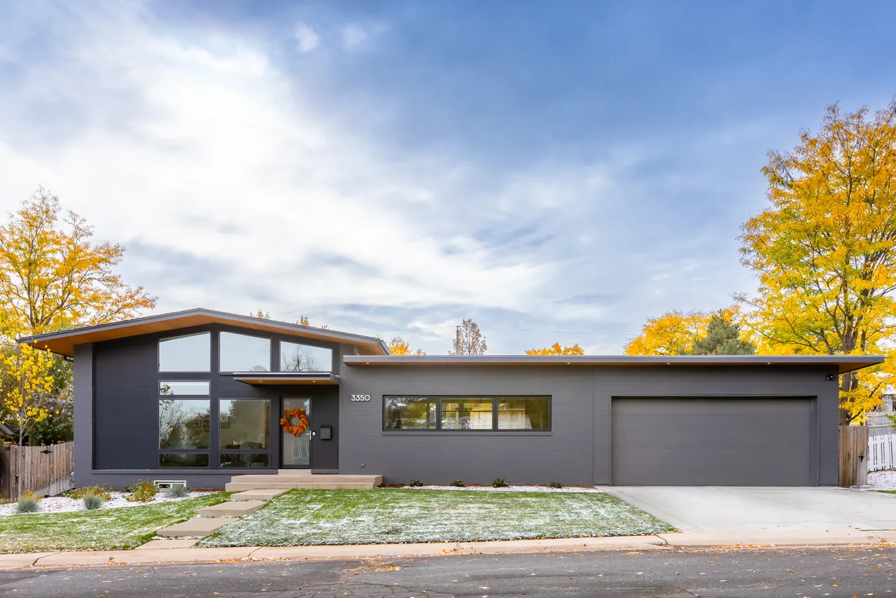Exterior view of a modern ranch-style house with dark siding and large front windows under a partly cloudy sky.