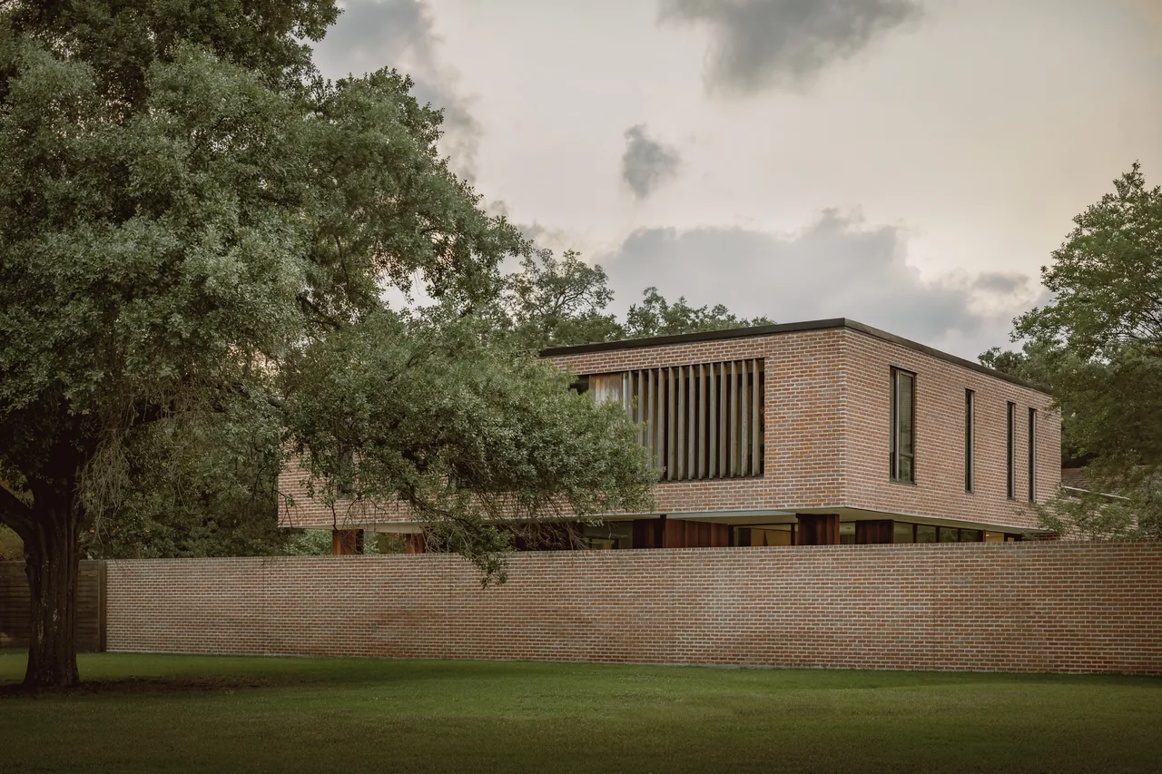 Two-story brick house with vertical wooden window slats and a brick privacy wall in a green yard under a cloudy sky.