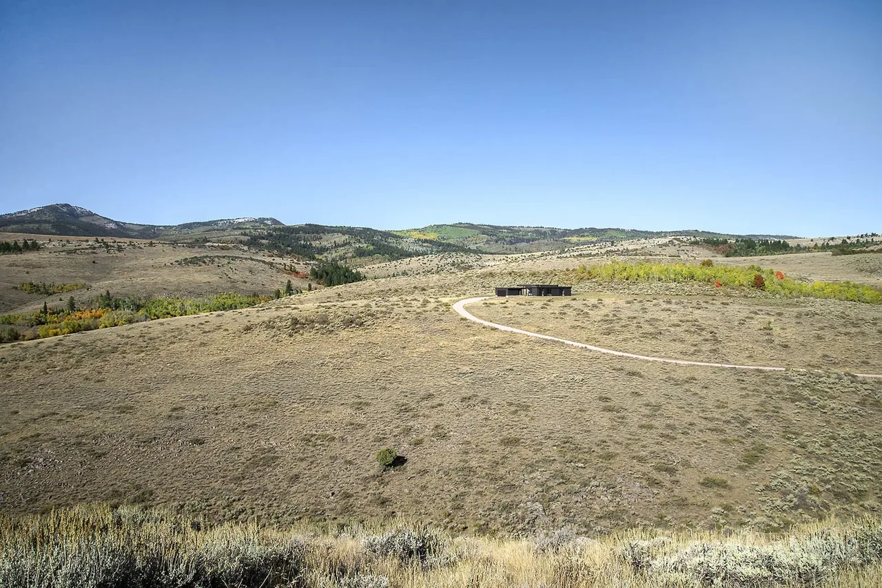 A single-story modern house sits isolated on a dry grassy hillside with a curved driveway under a clear blue sky.