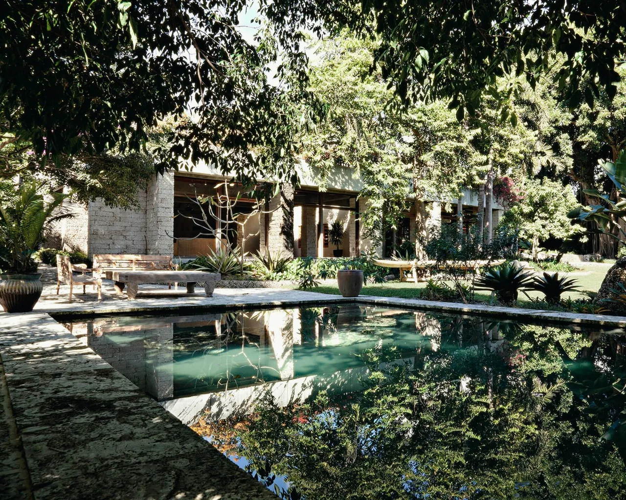 View of a residential garden with a reflecting pool surrounded by stone paving and lush greenery near a stone-columned house.