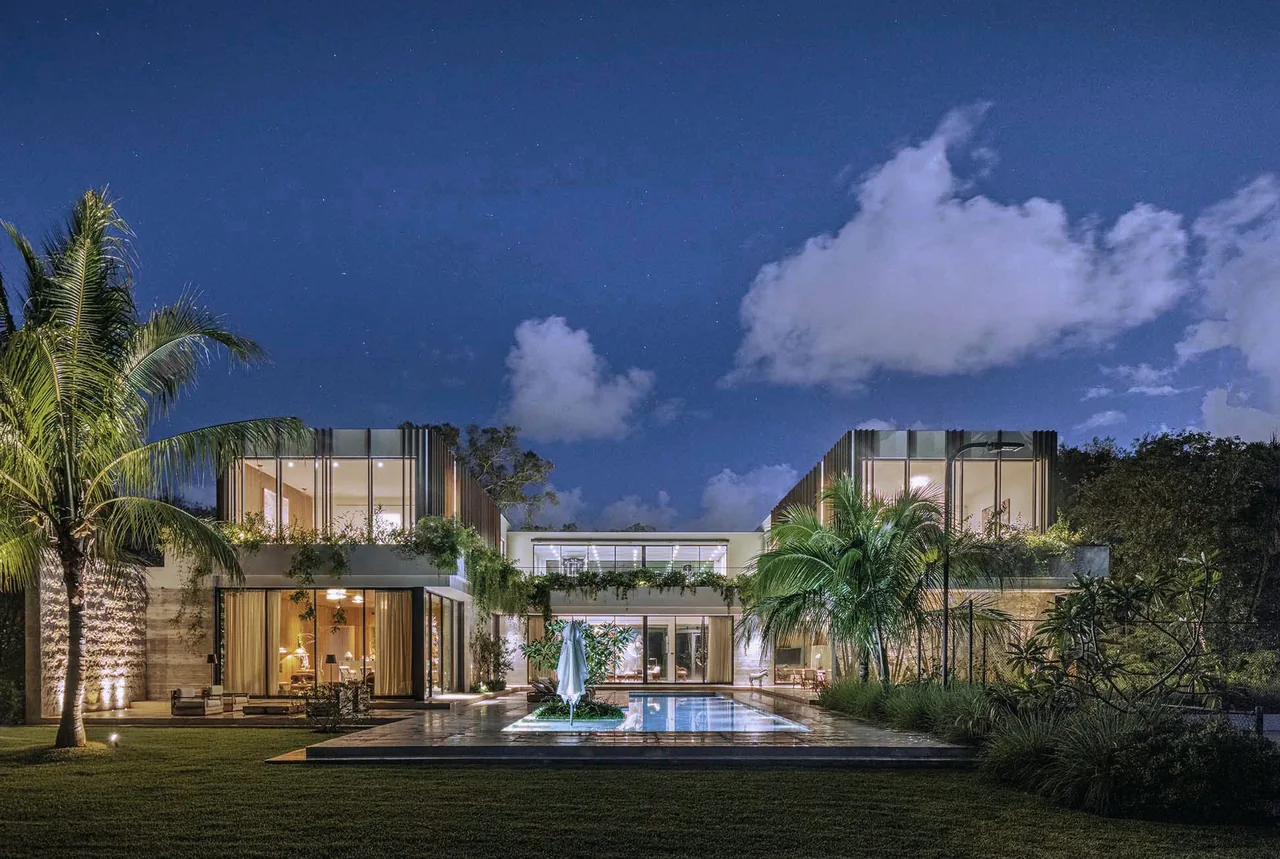 Night view of a modern two-story home with a pool and tropical landscaping under a cloudy sky.
