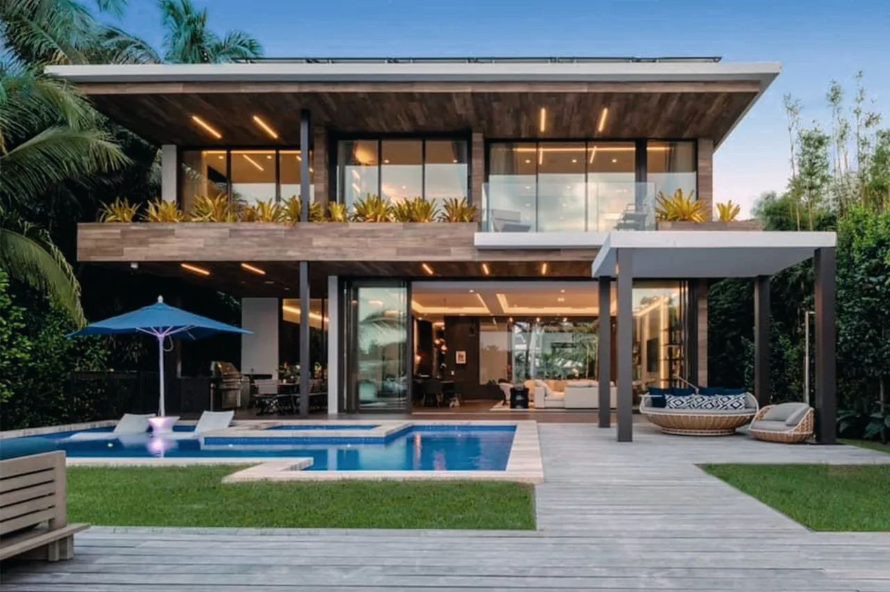 Two-story modern house with floor-to-ceiling glass walls overlooking a swimming pool and patio with outdoor furniture and an umbrella.