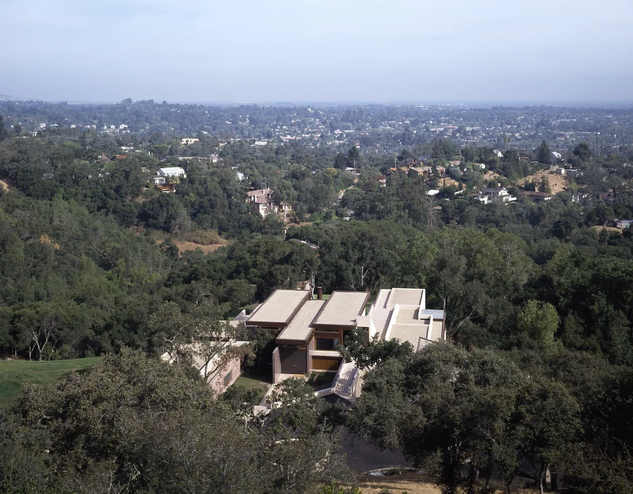 A modern house with flat roofs nestled among dense trees on a hillside overlooking a distant cityscape under a hazy sky.