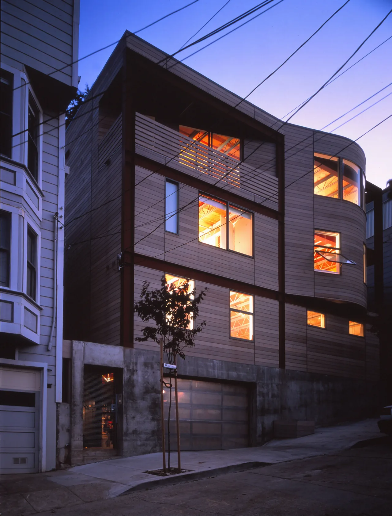 Three-story modern house with wood siding and glowing windows at dusk on a sloped street.