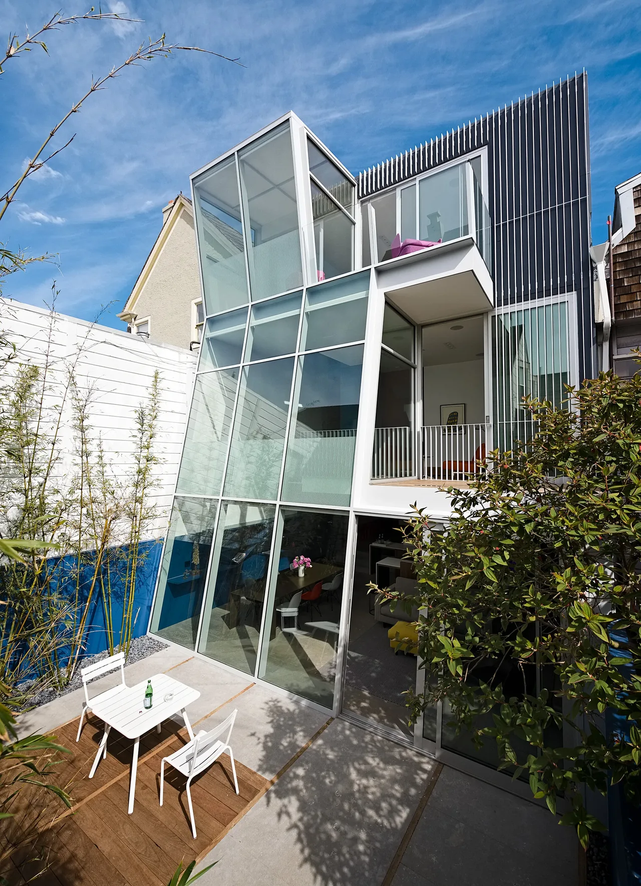 Rear view of a modern multi-level house with extensive floor-to-ceiling glass walls opening to a small courtyard with outdoor seating.