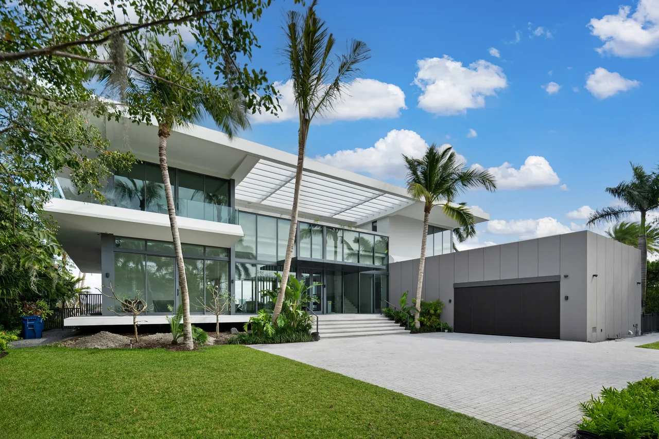 Modern two-story house with flat roof, large glass windows, and a spacious driveway under a blue sky.