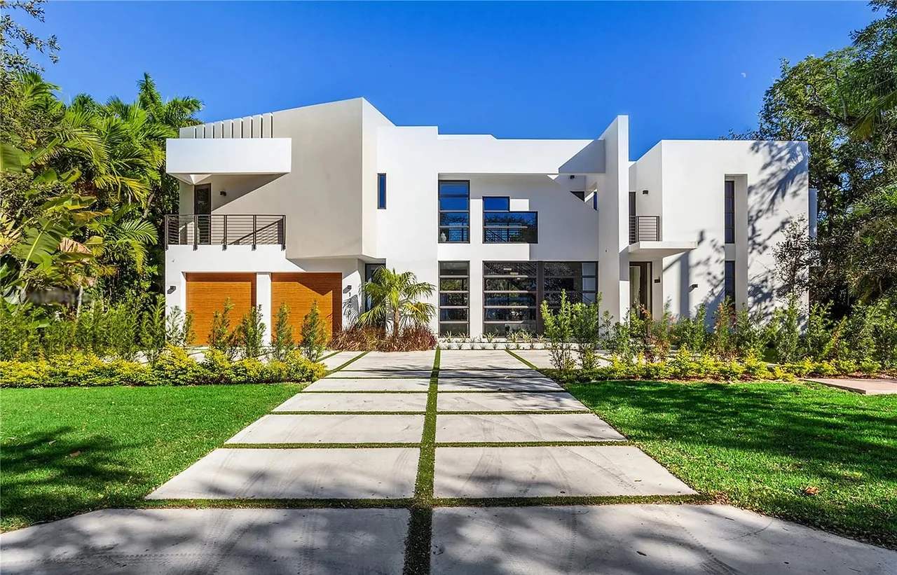 Front view of a modern white two-story house with large windows, metal balcony railings, and a driveway leading to wooden garage doors.