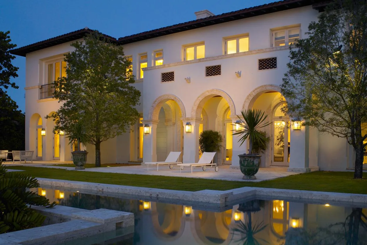 Evening view of a two-story white stucco house with arched outdoor hallways and a lit swimming pool in front.