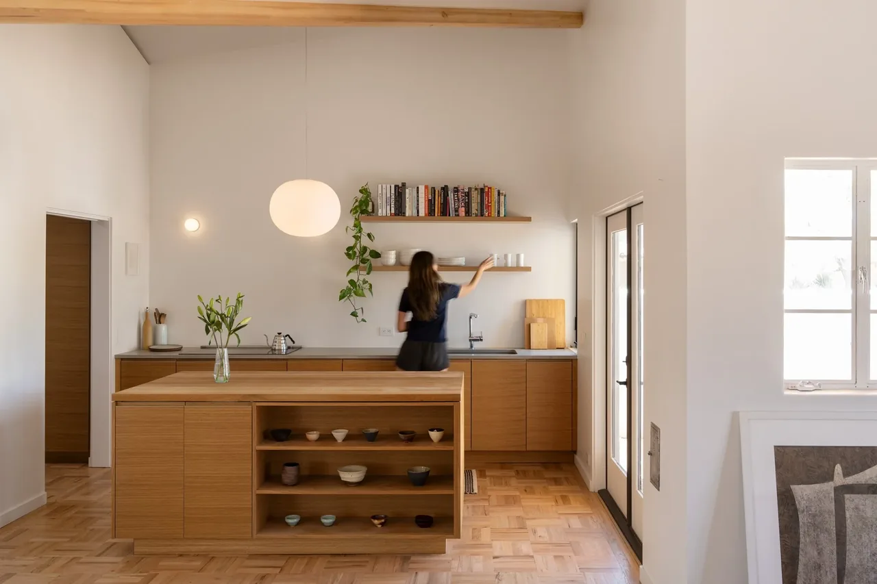 Interior view of a kitchen with a wooden island featuring open shelving, wooden cabinetry, and a woman reaching for dishes on floating wood-